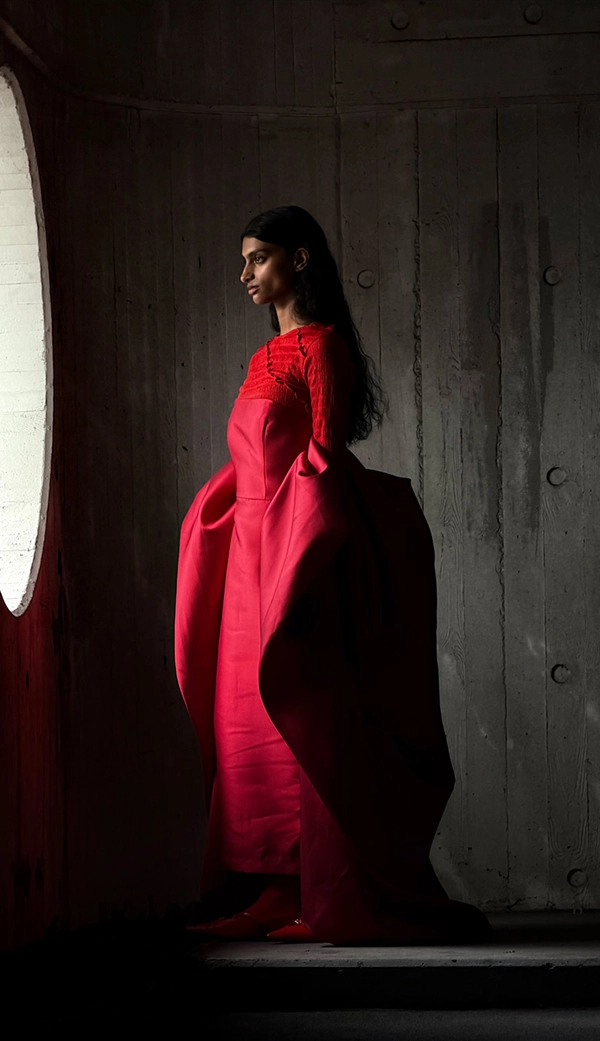 Женщина в длинном красном платье стоит в пустой комнате, пока ее фотографируют. Приложение «Фото» переключает фокусные расстояния, чтобы продемонстрировать возможности профессиональной камеры: 100 мм