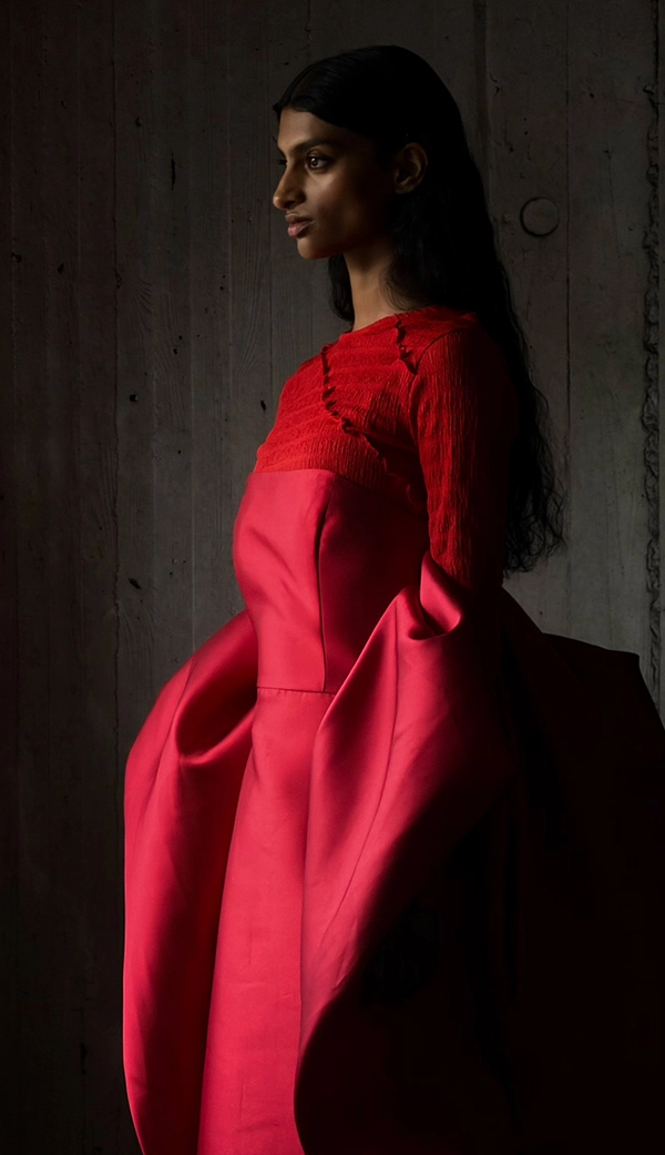 Женщина в длинном красном платье стоит в пустой комнате, пока ее фотографируют. Приложение «Фото» переключает фокусные расстояния, чтобы продемонстрировать возможности профессиональной камеры: 200 мм