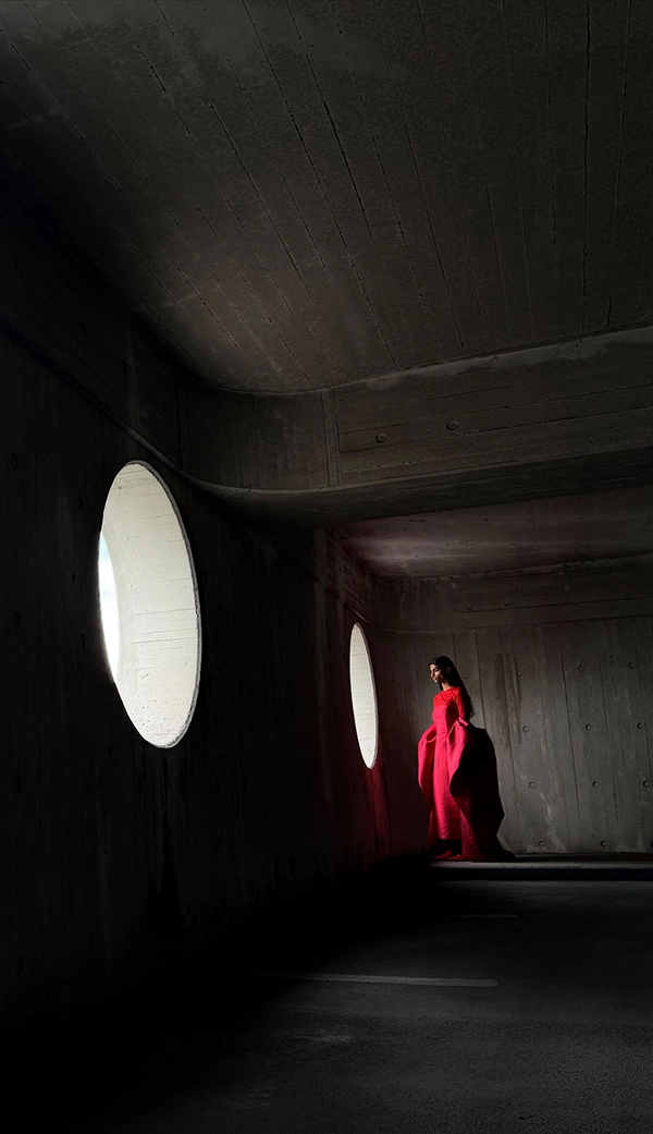 Женщина в длинном красном платье стоит в пустой комнате, пока ее фотографируют. Приложение «Фото» переключает фокусные расстояния, чтобы продемонстрировать возможности профессиональной камеры: 24 мм