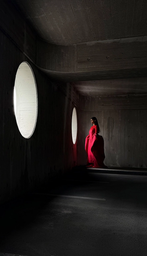 Женщина в длинном красном платье стоит в пустой комнате, пока ее фотографируют. Приложение «Фото» переключает фокусные расстояния, чтобы продемонстрировать возможности профессиональной камеры: 28 мм